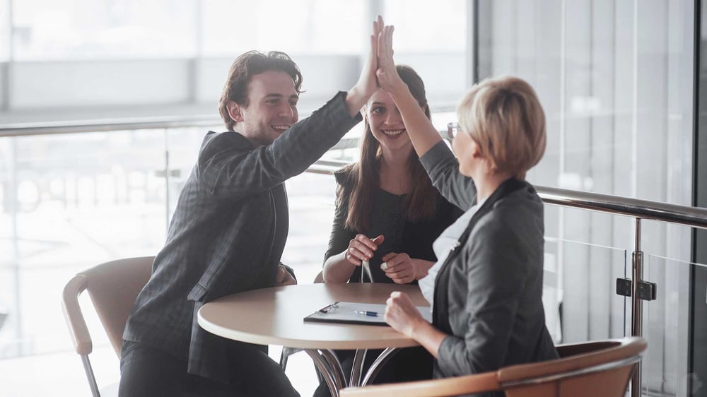 Eine Gruppe von drei Sales Leuten sitzt am Tisch und geben sich ein High Five